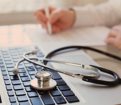 A stethoscope resting on a laptop's keyboard with a doctor writing a prescription in the background