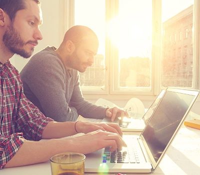 Two men each with a laptop sitting at a desk.