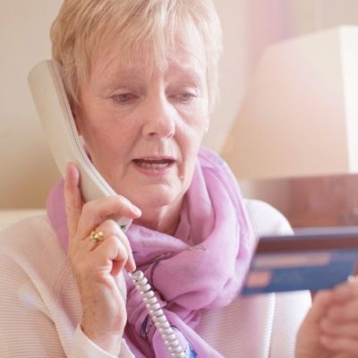 An elderly woman using a telephone while holding a credit card