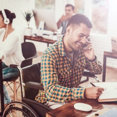 A man sitting in a wheelchair at a desk while using a cellphone and writing in a notebook