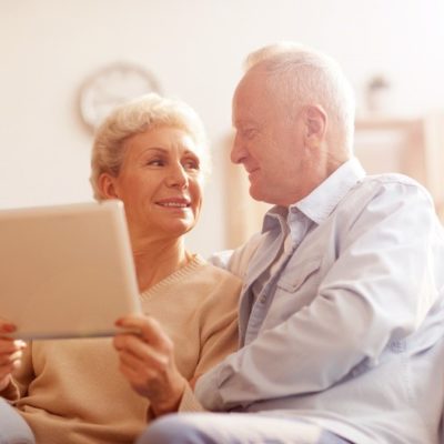 A couple smiling at one another while sitting on a couch holding a tablet device.