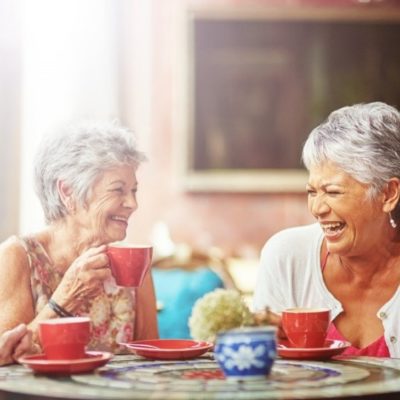 3 elderly women drinking coffee at a table while laughing