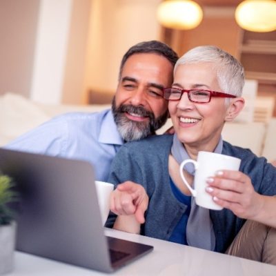 An older couple smiling and laughing while drinking coffee and using a laptop