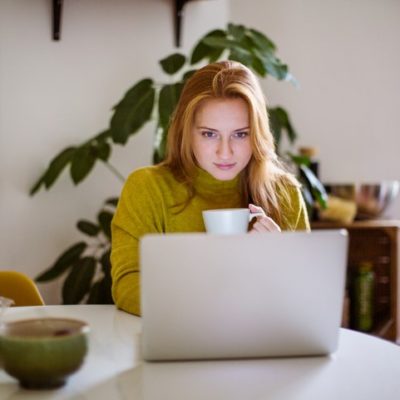 A woman sitting at a table using a laptop while holding a cup of coffee.