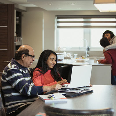 A grandfather and granddaughter are sitting at the kitchen table using a laptop whilst mother and son are hugging in the background.